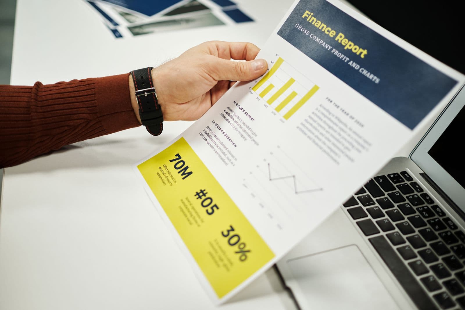 Une main tient un rapport financier imprimé à côté d'un ordinateur portable